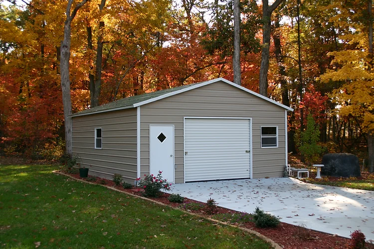 A Tan metal building with white doors and trim surrounded by a forest in the fall.